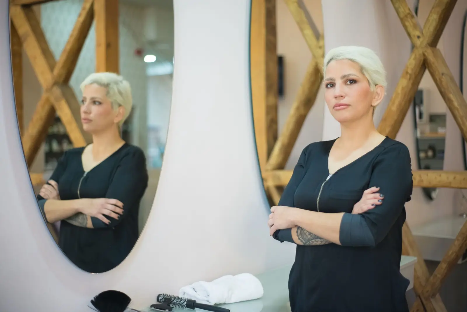 Ida, Friseurin mit kurzen hellen Haaren, steht mit verschränkten Armen vor dem Spiegel