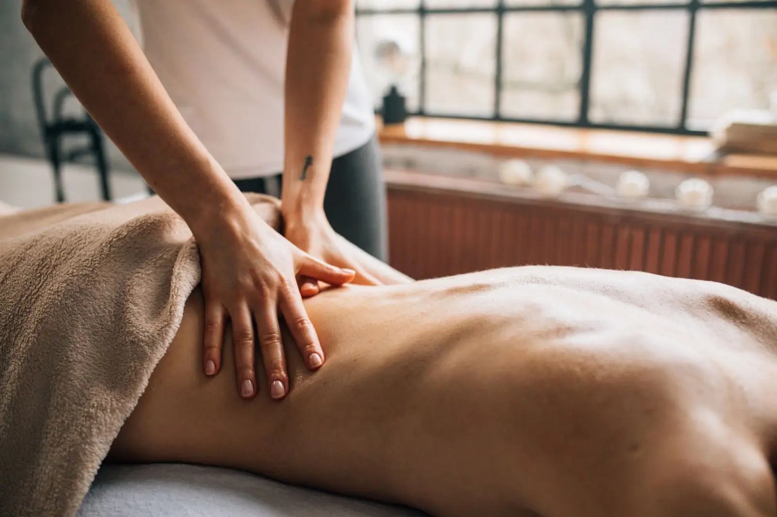 Masseurin massiert mit ruhigen Händen den Rücken einer Frau, Licht fällt durch große Fenster